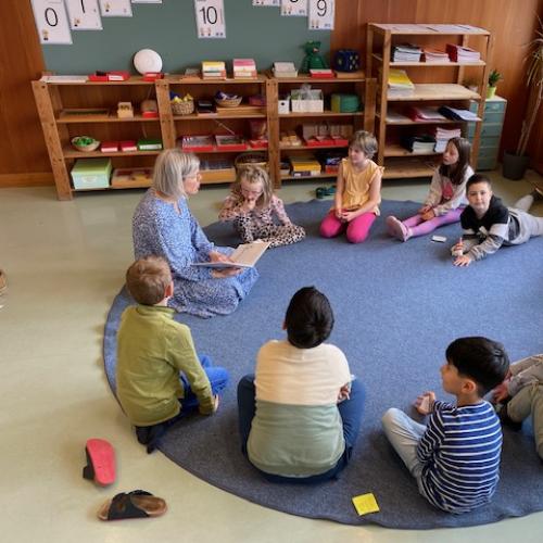 eine Gruppe Kinder sitzt am Boden auf einem blauen Teppich - die Lehrerin liest gerade vor