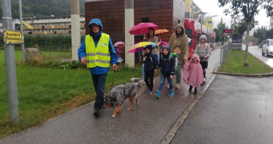 Mann mit gelber Warnweste und Hund geht einer Gruppe Kinder voraus am Gehsteig